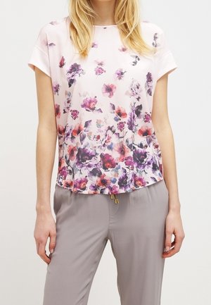 Vrouw draagt een wit blouse met korte mouwen, met paars-roze bloemenprint, gecombineerd met lichtgrijze broek, staand tegen een effen achtergrond.