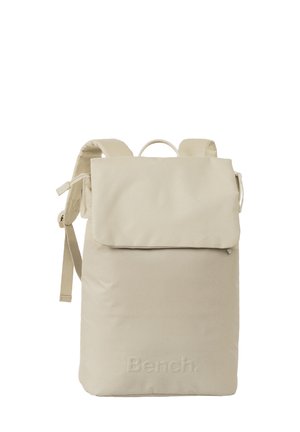Sac à dos beige avec un design à rabat, fabriqué en tissu. Il possède des bretelles ajustables, un logo à l'avant et une poignée de transport en haut.