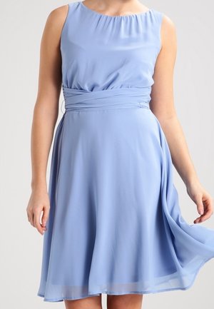 Femme portant une robe sans manches bleu clair longueur genou avec une taille froncée, tenant légèrement relevé le tissu de la robe sur un côté.