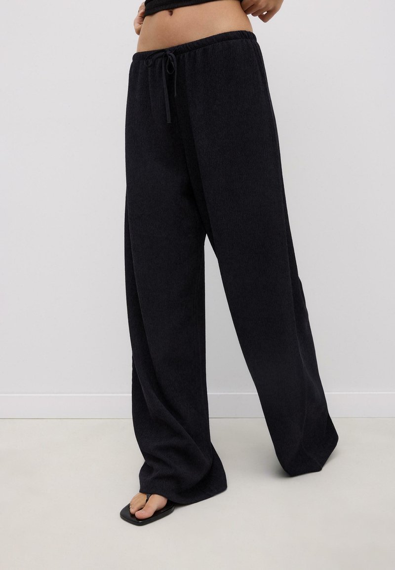 Femme portant un pantalon large noir texturé avec une ceinture élastique et un cordon de serrage, associée à des sandales noires à bout carré.