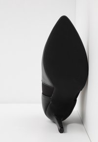 Chaussure à talon haut noire à bout pointu vue par la semelle avec un talon fin sur fond blanc.