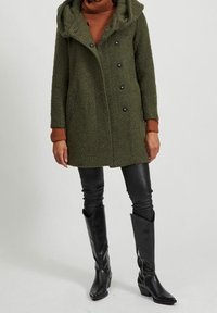Cappotto verde oliva, texture, con cappuccio e grandi bottoni, abbinato a un maglione a collo alto color ruggine e leggings in pelle nera lucida con stivali.