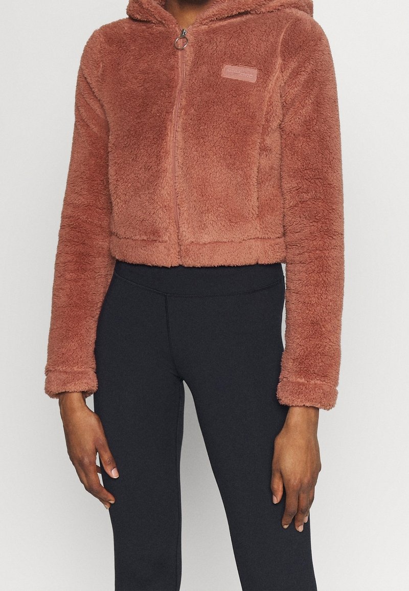 Chaqueta con capucha recortada en suave y texturizado forro polar coral. Cuenta con una cremallera frontal, un bolsillo de parche y puños elásticos. Combinada con leggins negros.
