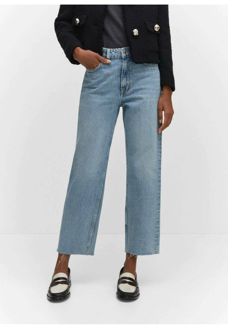 Personne portant un jean bleu clair court à jambes larges, une veste noire et des mocassins noirs et blancs, debout sur un fond blanc.