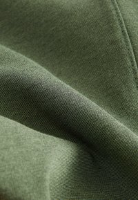 Tejido verde oliva con una superficie lisa y texturizada. El material presenta hilos finos y uniformes, así como variaciones sutiles en el tono.