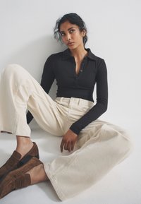 Black long-sleeve shirt with button placket, cream wide-leg pants, and brown suede shoes. Smooth textures, relaxed fit, and subtle design details.