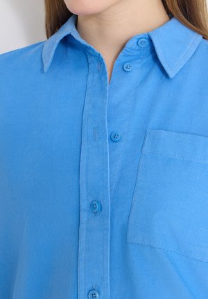 Nærbillede af en person iført en blå skjorte med knapper, krave og brystlomme, hvor knapper og stoftekstur er synlige.