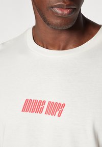 Tricou alb din bumbac, cu un logo roșu „ADIDAS HOOPS” curbat imprimi pe piept, având gât rotund și mâneci scurte.