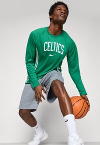 Giocatore di basket maschile con maglia a maniche lunghe verde dei Celtics, pantaloncini grigi, scarpe da ginnastica bianche, che palleggia una palla da basket arancione guardando verso l'alto.