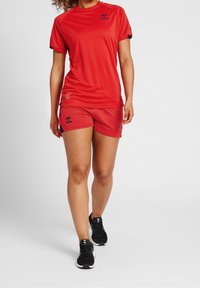 Camiseta deportiva roja y pantalones cortos a juego con logo, hechos de tejido suave. Zapatillas negras con detalles blancos. Mangas cortas y diseño ajustado.