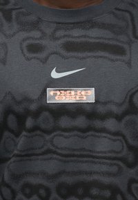 Σκούρο γκρι μπλουζάκι με υφασμένο σχέδιο, ανοιχτό γκρι λογότυπο Nike swoosh και πορτοκαλί ορθογώνιο ταμπελάκι με στυλιζαρισμένο κείμενο.