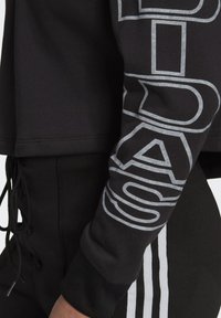 Svart sweatshirt med grå grafisk tryck på ärmen, med en avslappnad passform och ribbad muddar. Flätade dragsnören är synliga vid midjan.