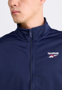 Námornícka zipová bunda vyrobená z hladkej tkaniny s vysokým golierom; obsahuje biele logo Reebok na hrudi a stredový zips.