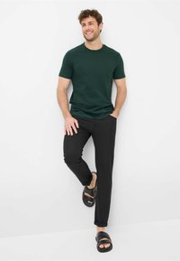 T-shirt verde scuro a maniche corte, pantaloni neri slim-fit e sandali neri a scivolo. Tessuto morbido e design casual. Modello in piedi su uno sfondo bianco.