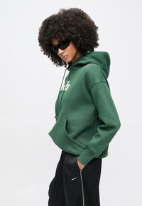 Zielona bluza z kapturem wykonana z miękkiego materiału, wyposażona w kieszeń kangurka oraz kaptur ze sznurkiem. Noszona z czarnymi spodniami dresowymi i okularami przeciwsłonecznymi.