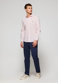 Camicia rosa chiaro a maniche lunghe con bottoni, abbinata a pantaloni blu scuro e sneakers bianche. Presenta un piccolo logo sulla camicia.