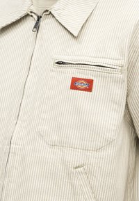 Veste beige et blanche à fines rayures avec fermeture éclair frontale, poche poitrine zippée et écusson logo Dickies rouge sur la poche poitrine.