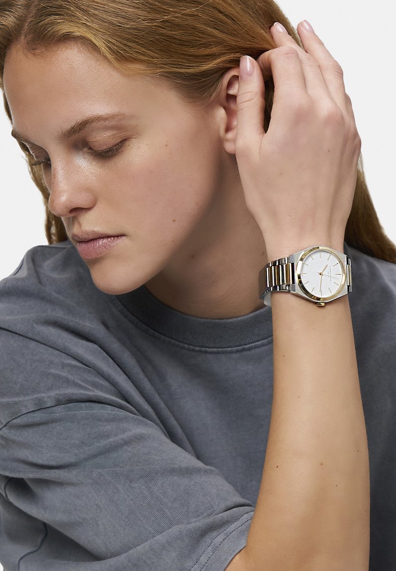Orologio argento e oro con quadrante rotondo bianco, cinturino in rete metallica e design minimalista, indossato su un polso. Maglietta grigia visibile.