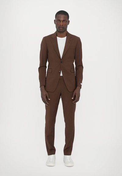 Brown tailored suit with single button closure and notch lapels, paired with a light gray t-shirt and white sneakers. Smooth fabric texture.