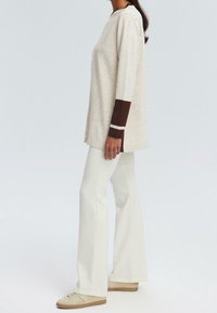 Maglia beige a maniche lunghe con accenti a righe marroni e bianche sulle maniche, abbinata a pantaloni a zampa d'elefante bianchi e sneakers beige.