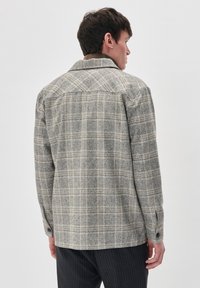 Jachetă cu carouri în nuanțe de gri și bej, realizată dintr-un material texturat. Are guler, manșete cu butoane și un design cu spate tip yoke, având o croială relaxată.
