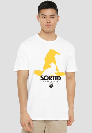 Young man wearing a white T-shirt with a yellow wizard hat, a white badger, and the text "Sorted Hufflepuff" on the front.