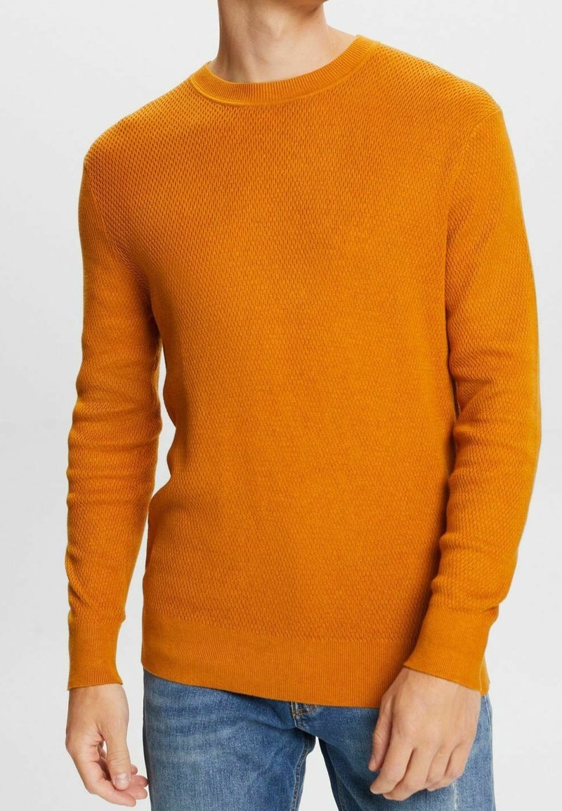 Homme portant un pull orange texturé à col rond et manches longues, ainsi qu'un jean bleu, debout devant un fond clair uni.