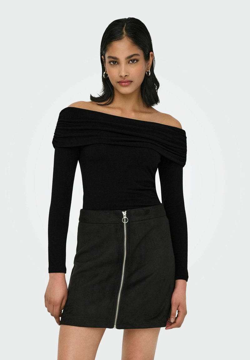 Top lungo nero con spalle scoperte e maniche lunghe, caratterizzato da pieghe a strati, abbinato a una minigonna in suede nera con chiusura a zip frontale.