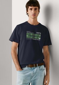 Jeune homme aux cheveux bruns courts portant un t-shirt bleu marine Pepe Jeans et un jean bleu clair, debout avec une main dans la poche.