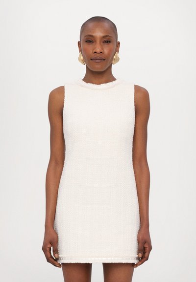 Teksturoidattu hihaton mekko off-white-värissä, korkealla kaula-aukolla ja repaleisilla reunoilla, mely ulottuu polven ylle. Kultasävyiset korvakorut täydentävät asua.