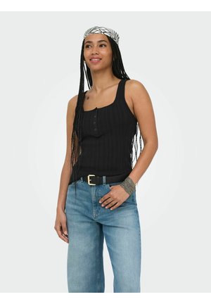 Ung kvinde med lange fletninger iført en sort ærmeløs top, blå jeans, et mønstret tørklæde og et sølvarmbånd, smilende og poserer.