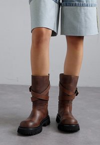 Bottines en cuir marron avec une finition texturée, présentant des sangles croisées et une semelle en caoutchouc noir épaisse.