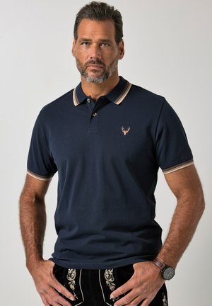Man with trimmed beard wearing navy polo shirt with striped collar and cuffs, embroidered deer logo, patterned black pants, and wristwatch.