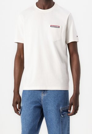 Mand iført en hvid T-shirt med korte ærmer og en brystlomme mærket "Hilfiger" og blå denim cargobukser.