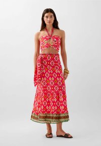 Top halter rosa con un davanti intrecciato, abbinato a una lunga gonna con motivo che presenta diamanti e bordi geometrici. Sandali marroni completano l'outfit.