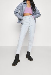 Kvinna poserar i ljusblå jeans, svarta snörstövlar, kort lila ribbad topp och grå läderjacka mot en enkel bakgrund.