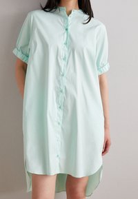 Robe chemise verte menthe à boutons, avec des manches courtes retroussées, présentant une coupe droite, un ourlet asymétrique et un tissu en coton doux.