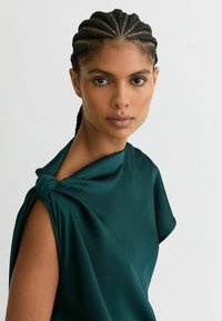 Tume-roheline satiintopp sõlmega õlal, millel on lahtine lõige ja sile tekstuur, neutraalse taustaga.