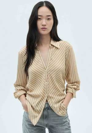 Camicia button-up beige a righe con colletto e maniche arrotolate, realizzata in tessuto leggero. Abbinata a jeans grigi.