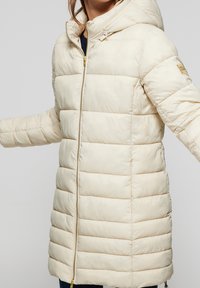 Veste rembourrée crème avec capuche, dotée d'une fermeture éclair intégrale et d'accents côtelés. Sa texture lisse et ses coutures horizontales ajoutent de la dimension.