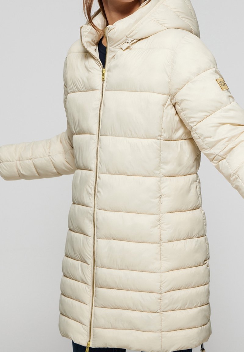 Veste rembourrée crème avec capuche, dotée d'une fermeture éclair intégrale et d'accents côtelés. Sa texture lisse et ses coutures horizontales ajoutent de la dimension.