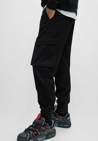 Pantalones cargo negros con bolsillos laterales, confeccionados con tejido resistente. Combinados con zapatillas deportivas negras y rojas que presentan detalles texturizados.