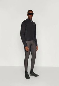 Modelo masculino vestindo uma jaqueta de ciclismo preta, calças de ciclismo cinzas, sapatos de ciclismo pretos e óculos de sol escuros, posando contra um fundo simples.