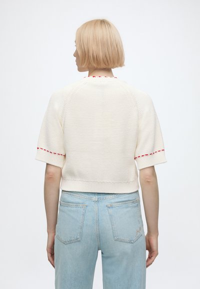 Personne aux cheveux blonds courts portant un pull blanc à manches courtes avec un liseré rouge et un jean bleu clair, vue de dos sur un fond uni.