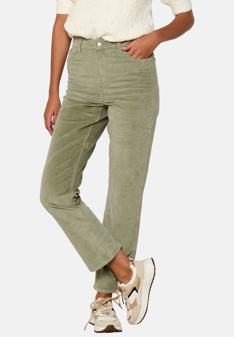 Personne portant un pantalon en velours côtelé vert clair, un pull texturé crème et des baskets blanches beige, debout avec une main dans la poche.