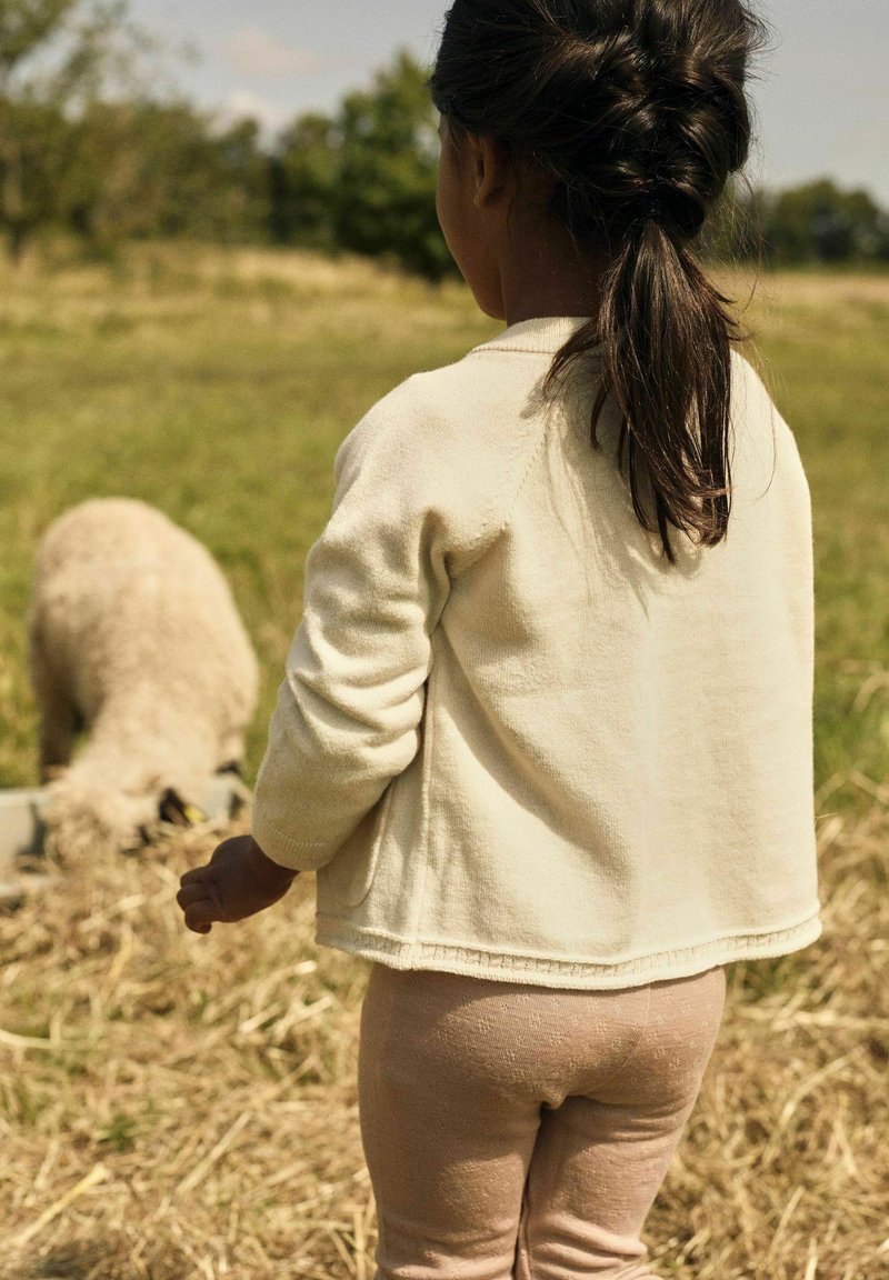 Un enfant portant un pull crème et des leggings roses pâles se tient dans un champ herbeux, faisant face à un mouton qui se nourrit à proximité.
