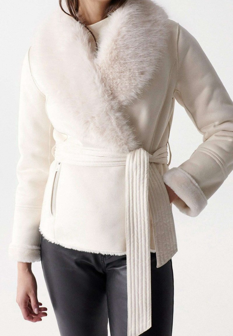 Cremefarbene Wickeljacke mit einem Kragen und Manschetten aus Kunstpelz, tailliert mit Gürtel und strukturierter Stoff. Verfügt über seitliche Reißverschlusstaschen und gerippte Akzente.