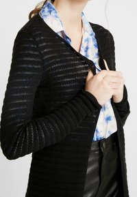 Černý pletený kardigán se strukturovaným vzorem a pruhy, s přiléhavým střihem a dlouhými rukávy, nošený přes tričko s modro-bílým tie-dye vzorem.