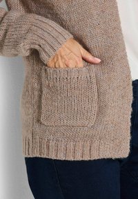 Hand in der Tasche eines beigen Strickpullovers, der über dunkelhosen getragen wird, zeigt die detaillierte Textur des Pulloverstoffs und den gerippten Saum.