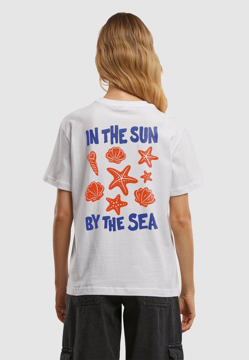 Personne aux longs cheveux portant un T-shirt blanc montrant des coquillages orange et des étoiles de mer avec un texte bleu « IN THE SUN BY THE SEA » dans le dos.